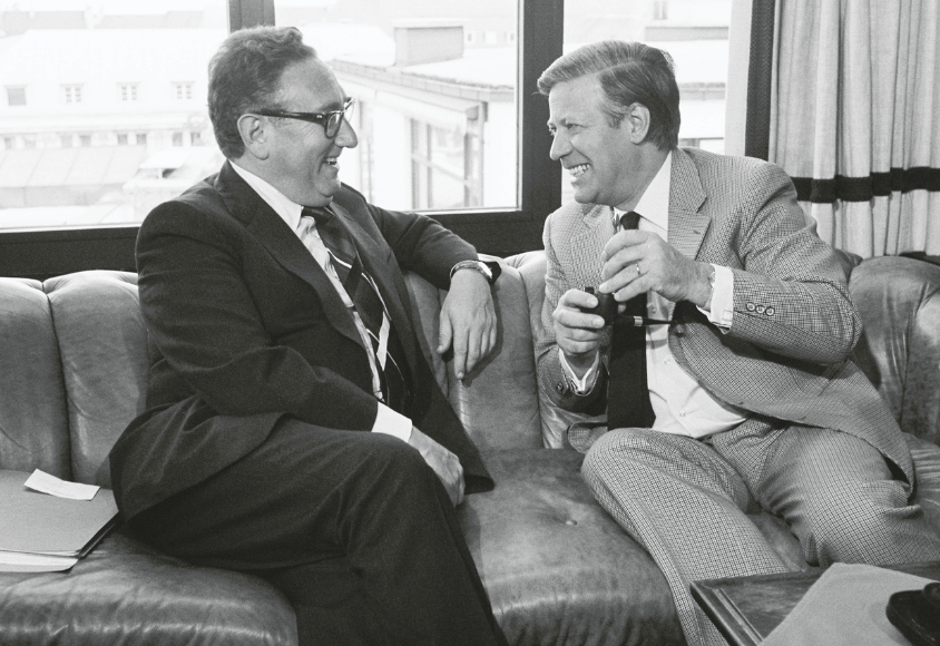 Schmidt und Kissinger sitzen lächelnd auf einem Ledersofa und unterhalten sich. Helmut Schmidt hält eine Pfeife.