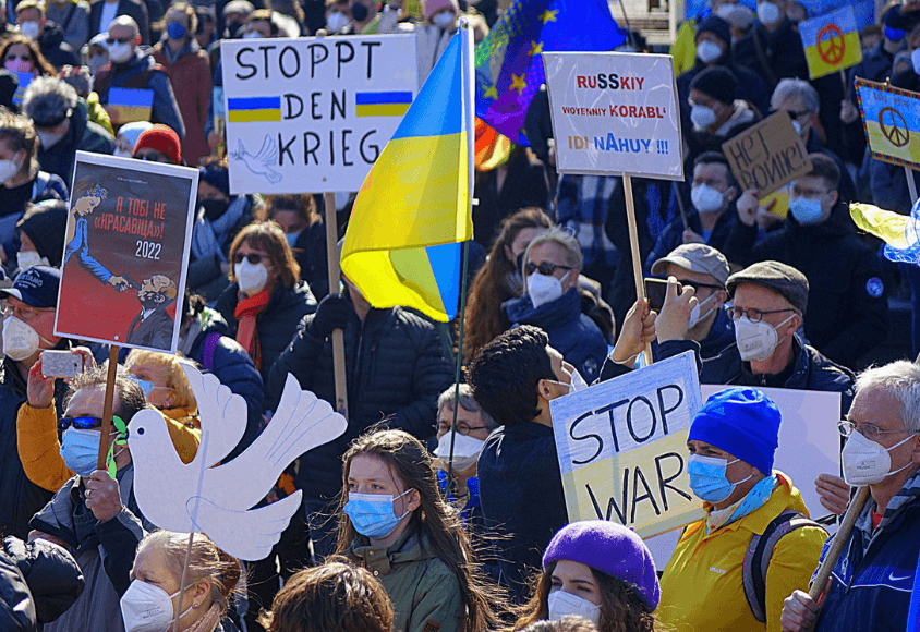 Demonstrierende halten Plakate hoch. Unter anderem mit der Aufschrift „Stoppt den Krieg“. Sie tragen medizinische Masken.
