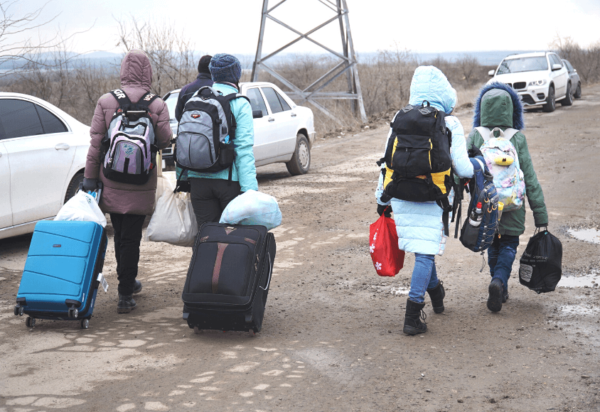 Erwachsene und Kinder laufen in Winterbekleidung, mit Koffern und Rucksäcken über eine matschige Straße.