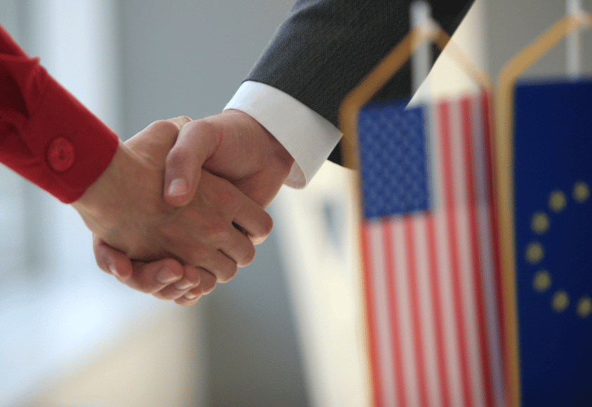 Nahaufnahme eines Händedrucks zwischen zwei Personen neben einer US-amerikanischen Flagge und europäischen Flagge.