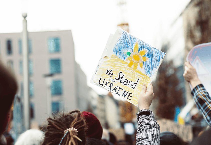 Eine Frau hält ein Schild auf einer Demonstration. Darauf ist die Ukrainische Flagge, eine gelbe Blüte und der Satz "We stand with Ukraine" abgebildet.