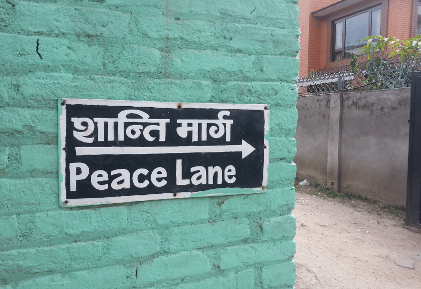 Auf einer hellblauen Wand hängt ein Schild mit der Aufschrift "Peace Lane" und einem Pfeil, der in die rechts abgehende Straße zeigt.