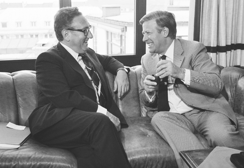 Henry Kissinger und Helmut Schmidt sitzen lachend nebeneinander auf einem Sofa. Das Bild ist eine historische Aufnahme und somit schwarz-weiß gehalten.