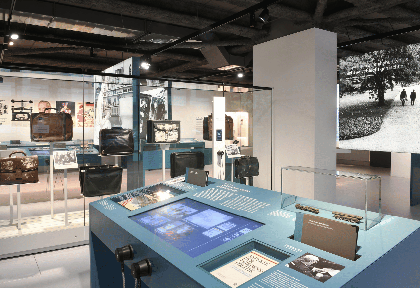 Ein blauer Tisch mit Monitor steht vor einer Vitrine mit vielen Arbeitstaschen. Im Hintergrund hängt ein großes Foto. Eine Aufnahme aus der Ausstellung "Schmidt! Demokratie leben"