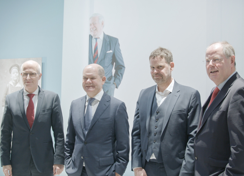 Vier Männer im Anzug stehen für ein Foto bereit: Peter Tschentscher und Per Steinbrück mit SPD-roter Krawatte, Olaf Scholz mit blaugrauer Krawatte und Magnus Koch in Weste unter dem Sakko. 