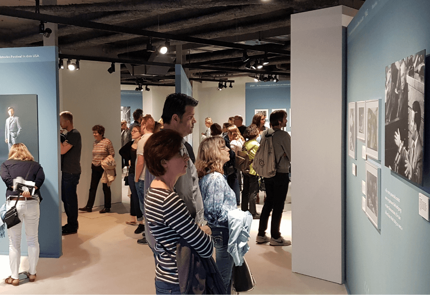 Viele Menschen laufen durch eine Ausstellung und schauen sich Fotos an. 