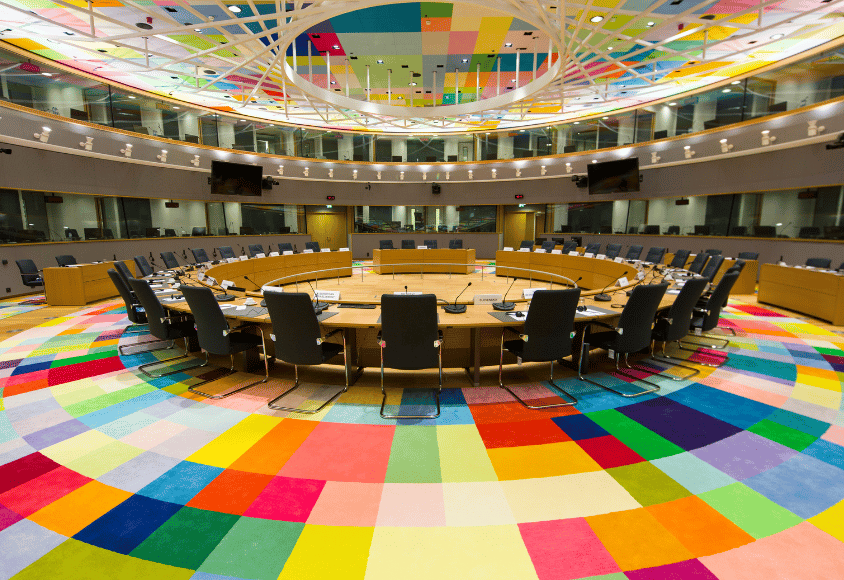 Ein runder Sitzungssaal mit einem runden Tisch in der Mitte und zahlreichen Stühlen. Der Teppich und die Decke sind mit bunten Quadraten übersäht.