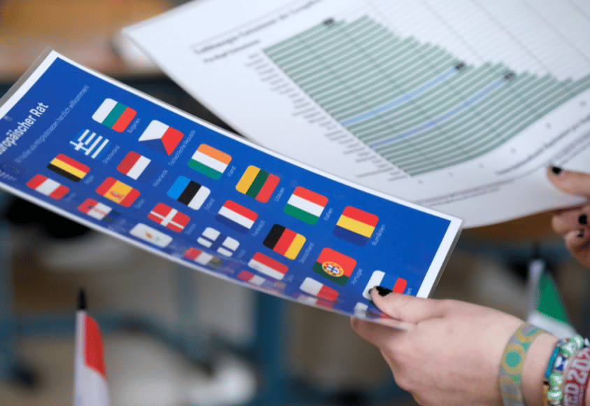 Zwei laminierte Zettel werden von einer Person gehalten. Darauf sind die Flaggen der Mitglieder des Europäischen Rats und eine verschwommene Tabelle zu sehen.