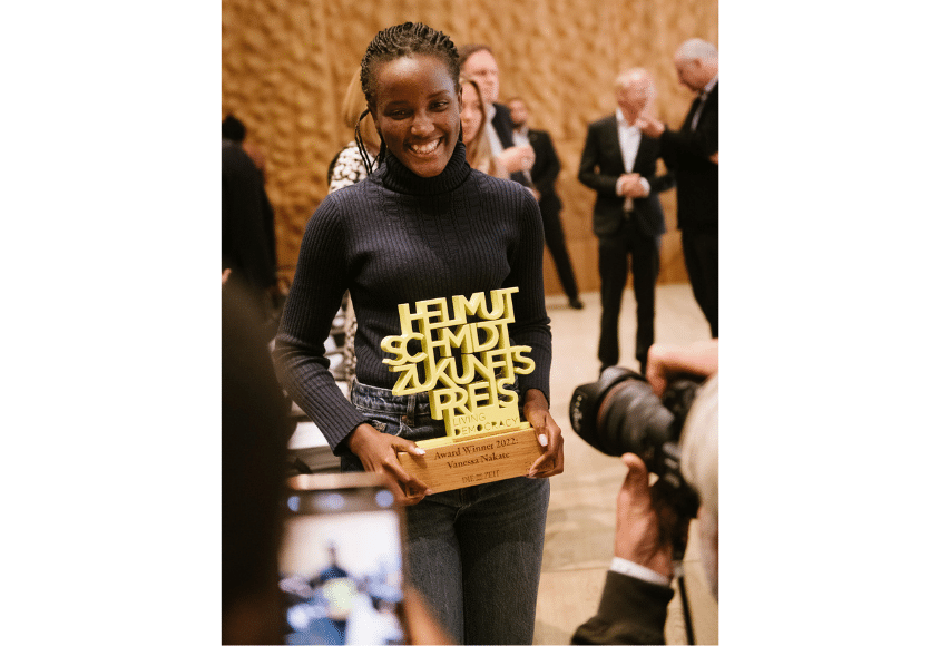 [Translate to English:] Vanessa Nakate in schwarzem Pullover mit dem gelben Award in der Hand. Sie lächelt.