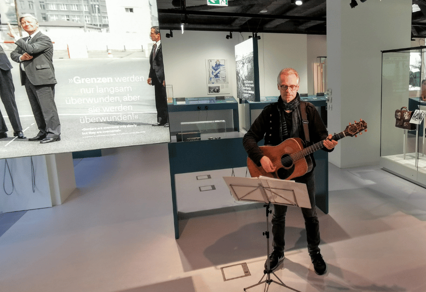 Mike Manske steht mit Gitarre und Notenheft in der Ausstellung "Schmidt! Demokratie leben".