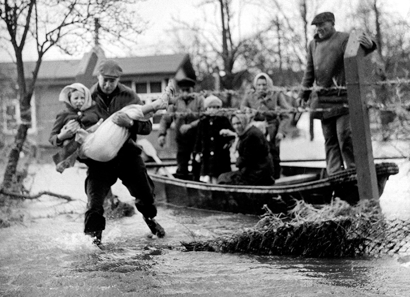 Momentaufnahme während der Hamburger Sturmflut. Ein Mann trägt ein Kind durch die wadenhohen Wassermassen. Im Hintergrund sind Menschen auf einem kleinen Boot.
