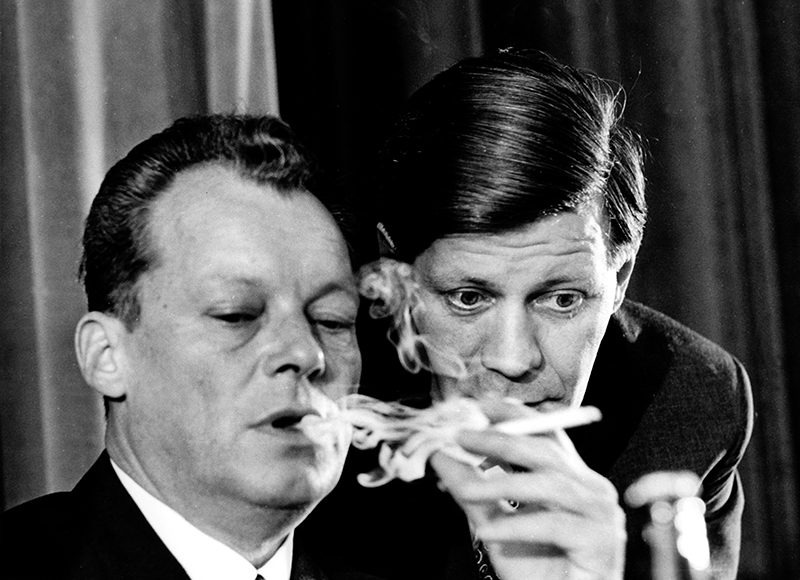 Willy Brandt raucht eine Zigarette. Helmut Schmidt steht hinter Brandt und lehnt sich zu ihm herunter.