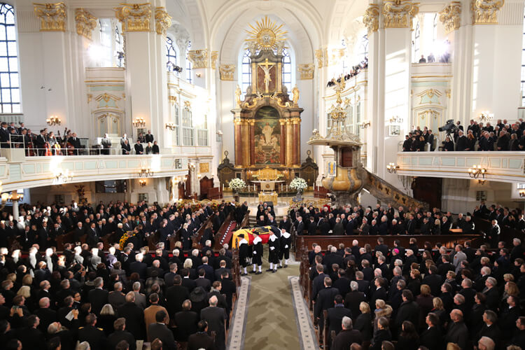 Ein Sarg bedeckt mit Deutschlandflagge wird durch eine Kirche mit Trauergästen getragen.