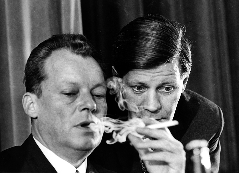 Helmut Schmidt beugt sich im Gespräch zu dem rauchenden Willy Brandt herunter.