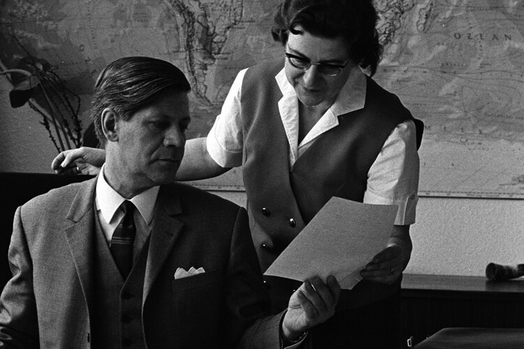 Eine Frau in Business-Kleidung zeigt Schmidt ein Dokument. 