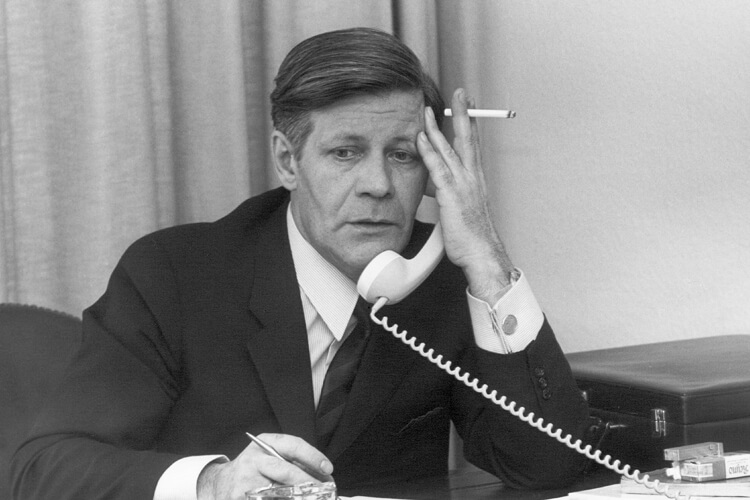 Helmut Schmidt hält einen Telefonhörer an sein Ohr. In der gleichen Hand hält er eine angezündete Zigarette.