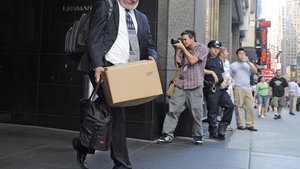 Man in suit with cardboard box in hand on Wallstreet, in the background photographer and policewoman