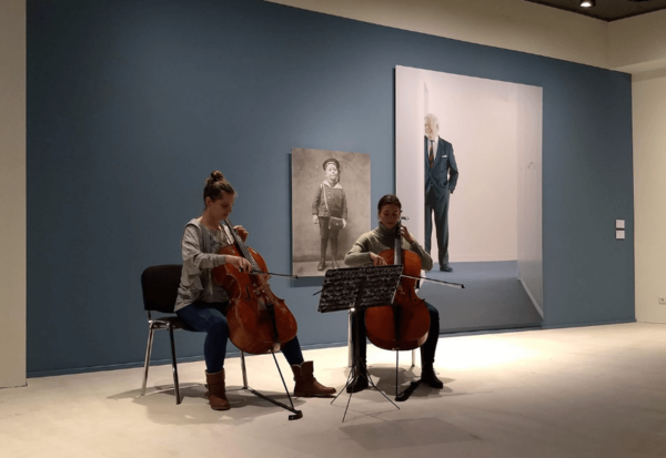 Zwei Cello spielende Frauen sitzen vor Fotos von Helmut Schmidt, die an einer Wand hängen.