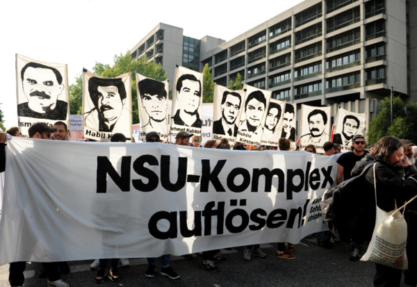 Demonstrierende halten Illustrationen der Opfer von NSU-Verbrechen und ein großes Tuch mit der Aufschrift „NSU-Komplex auflösen!“ hoch.