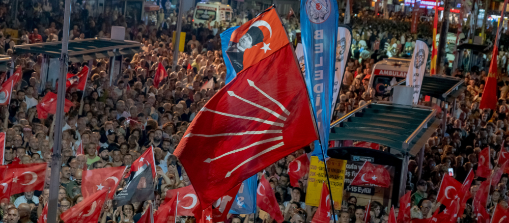 A crowd at a political rally, with many Turkish flags and red party banners fluttering above the crowd