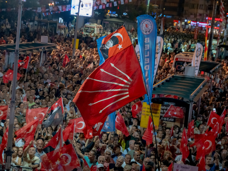 A crowd at a political rally, with many Turkish flags and red party banners fluttering above the crowd