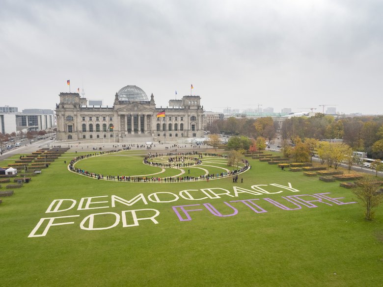 Teilnehmer des ersten Bürgerrates als lebendiges Kunstwerk in einer Spirale inszeniert auf der Wiese vor dem Reichstag. Auf dem Rasen neben ihnen liegt der Schriftzug "Democracy For Future".