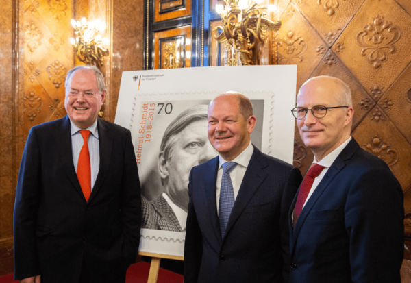 Drei Männer stehen vor einem Plakat, das eine Briefmarke mit Schmidts Gesicht zeigt.