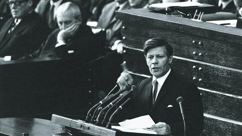 Black-and-white photograph, 1969: Helmut Schmidt is speaking at the lectern in the Bundestag, raising his finger; members of parliament are seated behind him. He is taking part in the debate on emergency legislation in the Bundestag.