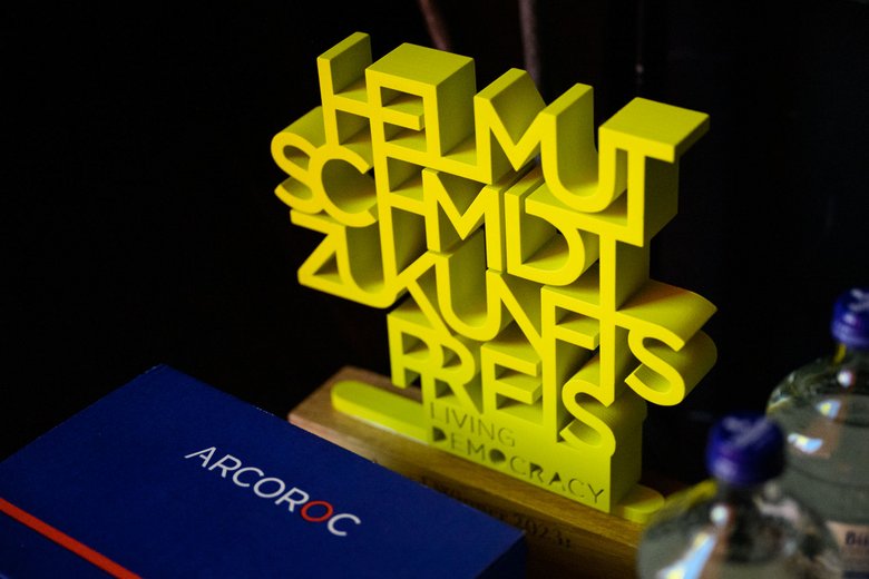 [Translate to English:] Award aus gelben Kunststoffbuchstaben mit dem Schriftzug "Helmut Schmidt Zukunftspreis - living democracy" auf einem Holzsockel. Der Helmut Schmidt Zukunftspreis 2023