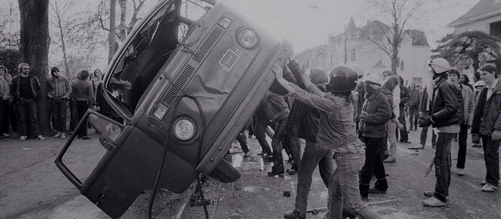 Schwarz-weiß Foto von Protestanten, die einen VW-Bus umstürzen