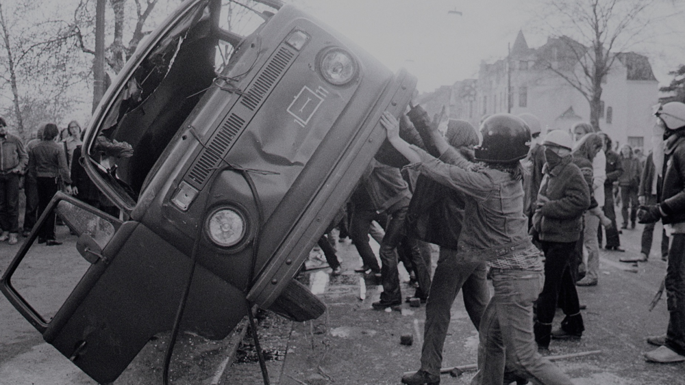 Schwarz-weiß Foto von Protestanten, die einen VW-Bus umstürzen