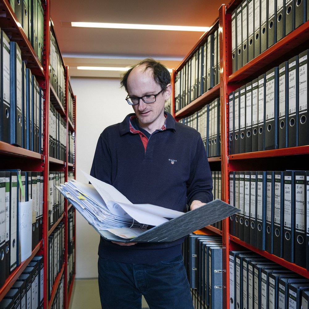 Man steht mit Zeitung in der Hand vor Archivschränken