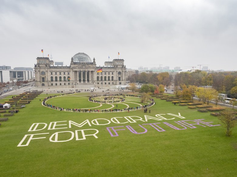 Participants in the first Citizens’ Assembly are arranged in a spiral on the lawn in front of the Reichstag, forming a living work of art. The words “Democracy For Future” are written on the grass beside them.