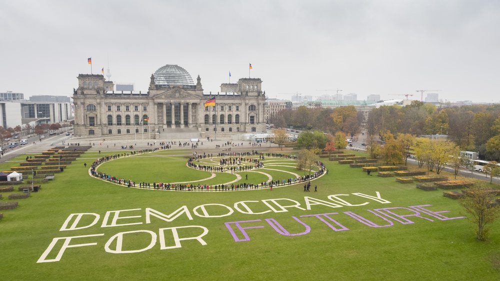 Teilnehmer des ersten Bürgerrates als lebendiges Kunstwerk in einer Spirale inszeniert auf der Wiese vor dem Reichstag. Auf dem Rasen neben ihnen liegt der Schriftzug "Democracy For Future".