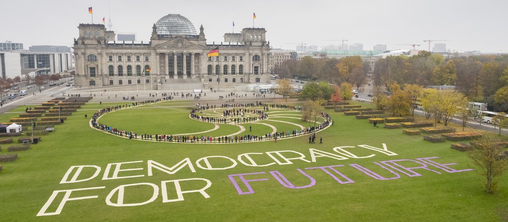 Teilnehmer des ersten Bürgerrates als lebendiges Kunstwerk in einer Spirale inszeniert auf der Wiese vor dem Reichstag. Auf dem Rasen neben ihnen liegt der Schriftzug „Democracy For Future“