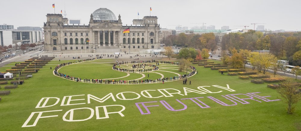 Participants in the first Citizens’ Assembly are arranged in a spiral on the lawn in front of the Reichstag, forming a living work of art. The words “Democracy For Future” are written on the grass beside them.