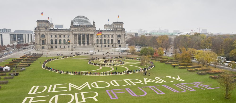 Teilnehmer des ersten Bürgerrates als lebendiges Kunstwerk in einer Spirale inszeniert auf der Wiese vor dem Reichstag. Auf dem Rasen neben ihnen liegt der Schriftzug "Democracy For Future".