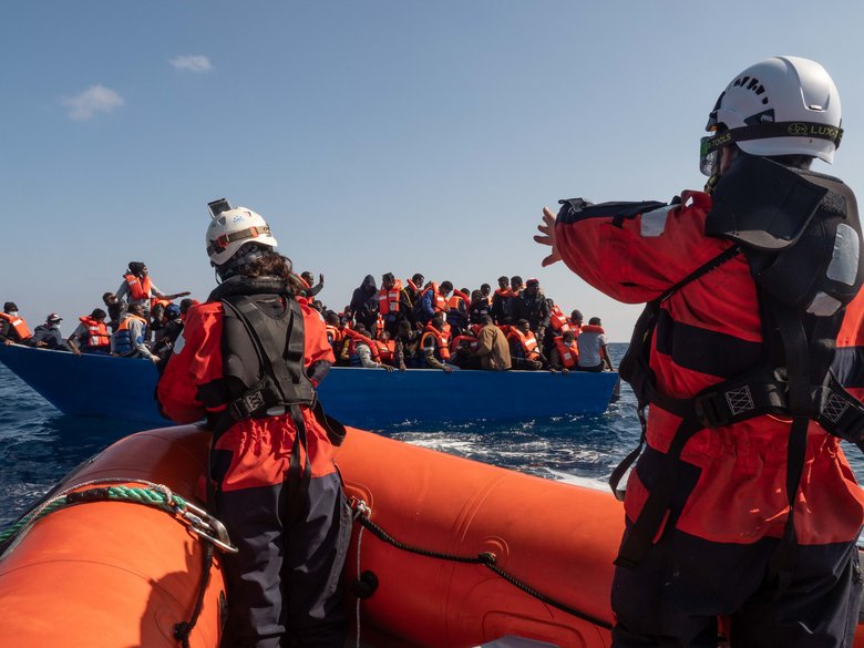 Rotes Boot der Sea-Watch mit zwei Personen vor blauem Boot mit zahlreichen Menschen auf Meer