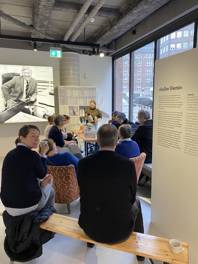 A small group of people are sitting together drinking coffee. A woman pours herself a new coffee at the coffee counter.