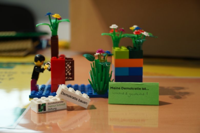 Colour photo: Several Lego bricks, including some shaped like flowers. One of the bricks bears the inscription: My democracy is growing and thriving.