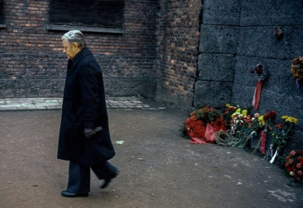 Helmut Schmidt läuft in schwarzer Kleidung und gesenktem Blick vor einer Mauer, an der Blumenkränze angelehnt sind.