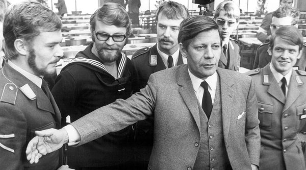 Helmut Schmidt, der seinen Arm vor mehreren uniformierten Männern ausstreckt.  