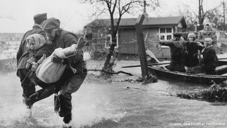 Ein Mann trägt ein Kind in knietiefem Wasser. Rechts daneben ein Boot mit drei Menschen.