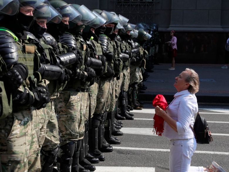 Demonstrator dressed in white with a red scarf in her hand kneels in front of a row of police officers