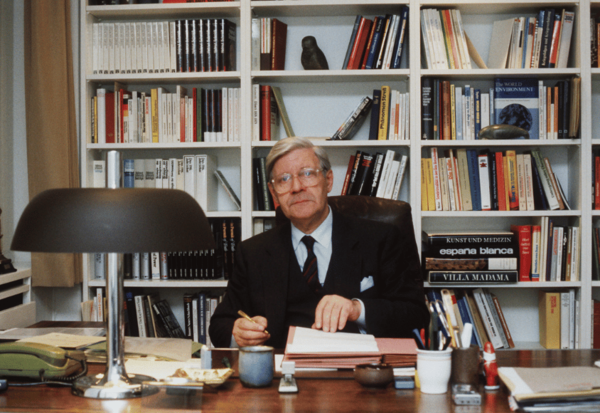 Helmut Schmidt sitzt am Schreibtisch und unterschreibt Dokumente. Im Hintergrund steht ein gut gefülltes Bücherregal.