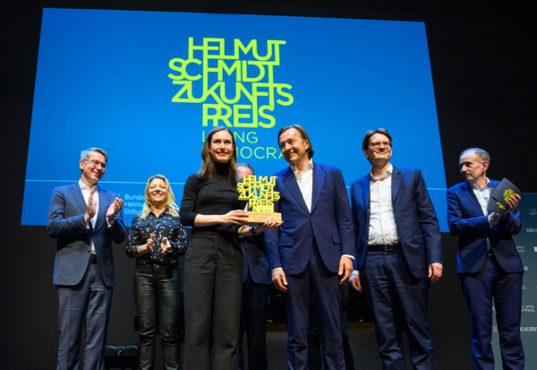 Sanna Marin hält den Helmut Schmidt Zukunftspreis. Auf der Bühne steht die Moderatorin und die Jury und applaudiert.