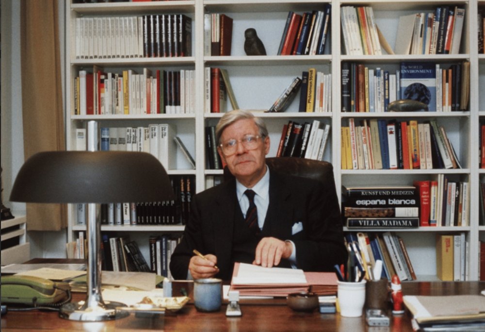 Helmut Schmidt sitting at a desk in front of a wall cupboard with books