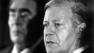 Black-and-white photograph: Helmut Schmidt in a suit, seen in profile; Soviet leader Leonid Brezhnev can be seen out of focus in the background.