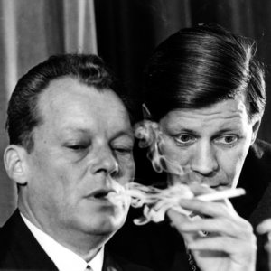Black-and-white photograph: Willy Brandt on the left, smoking a cigarette, with Helmut Schmidt to his right. The party executive, the Control Commission, the Party Council and the Social Democrats’ new parliamentary group in the Bundestag met for a joint session in Bad Godesberg on 25 September 1965.
