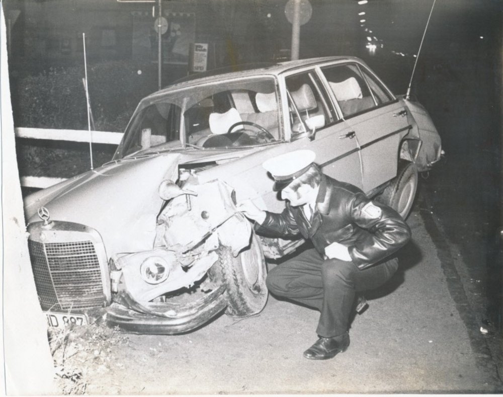 Black and white photograph Police officer kneels in front of demolished car
